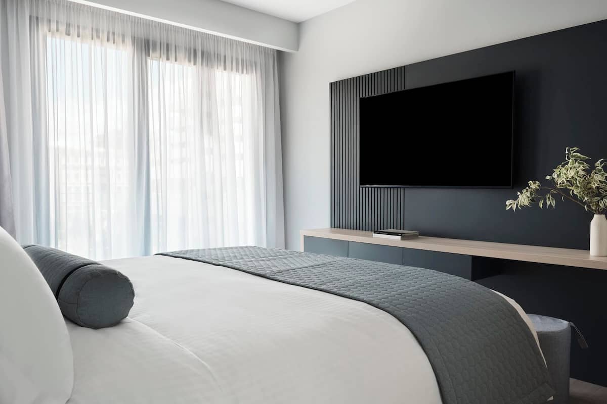 Un dormitorio moderno con una cama, un televisor montado en la pared y una ventana con cortinas translúcidas.