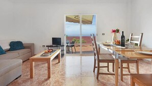Ein Wohnzimmer mit einer Couch, einem Couchtisch und einem Esstisch mit Stühlen. Ein Fernseher steht auf einem Schrank, und eine Glastür öffnet sich zu einem Außenbereich mit Meerblick.