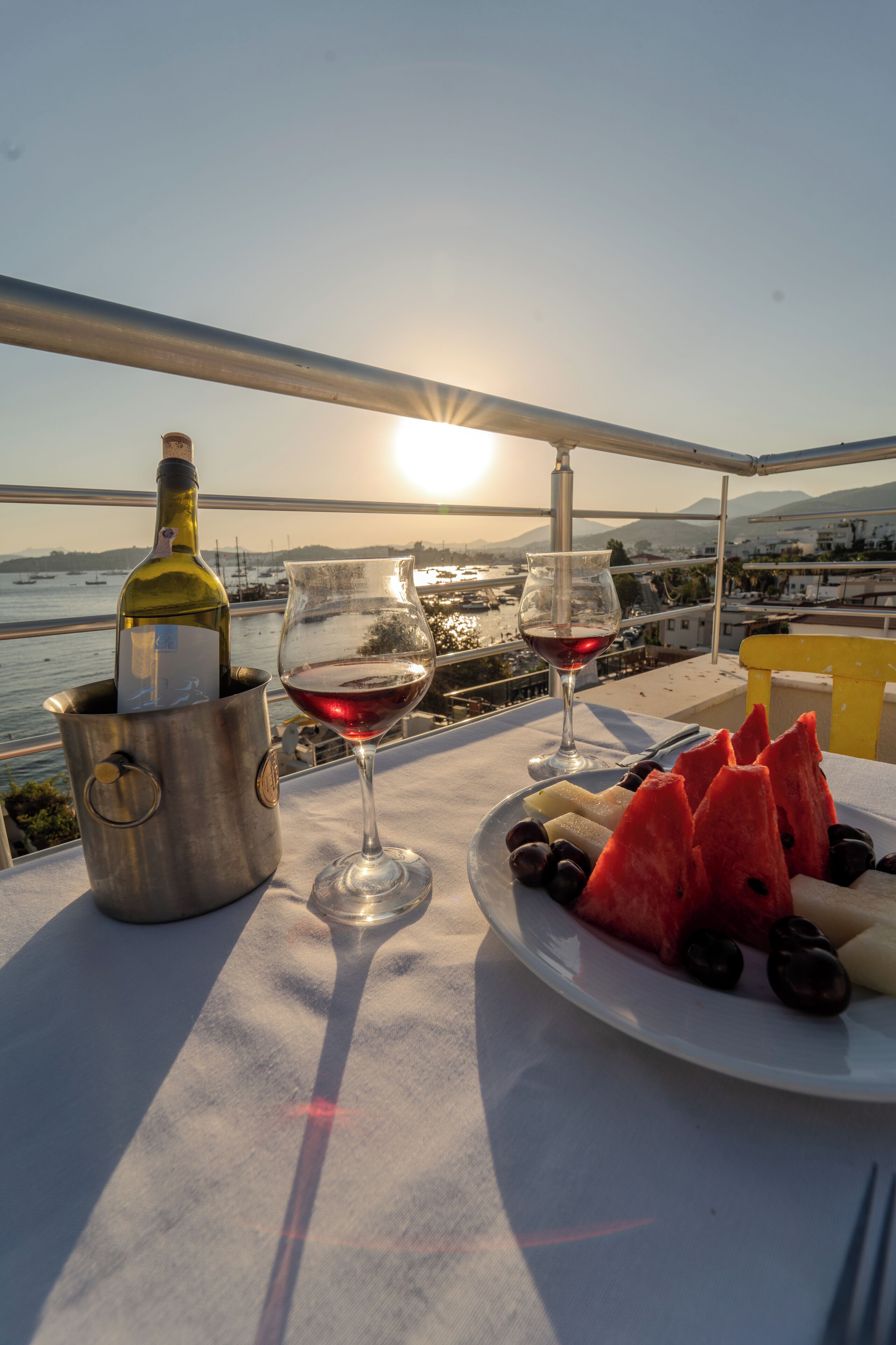 Une table avec une bouteille de vin, deux verres à vin et une assiette de pastèque et d’olives, sur un balcon donnant sur une étendue d’eau et des bateaux.