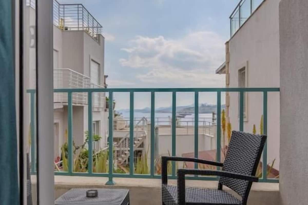 Un balcón con una silla y una mesita, vista a edificios y un cielo nublado.