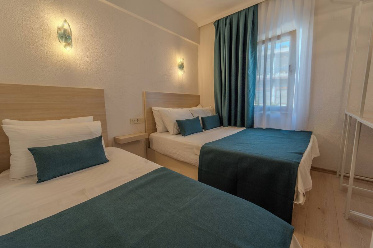 Habitación de hotel con dos camas, cada una con cabecera de madera, ropa de cama blanca y almohadones turquesa. Hay lámparas fijadas a la pared y una ventana con cortinas translúcidas.