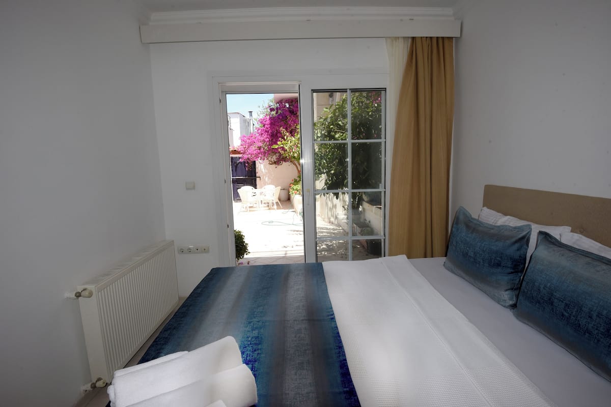 Un dormitorio con una cama grande, un radiador, y un ventanal corredero que da a un espacio exterior con una silla y una planta en maceta.