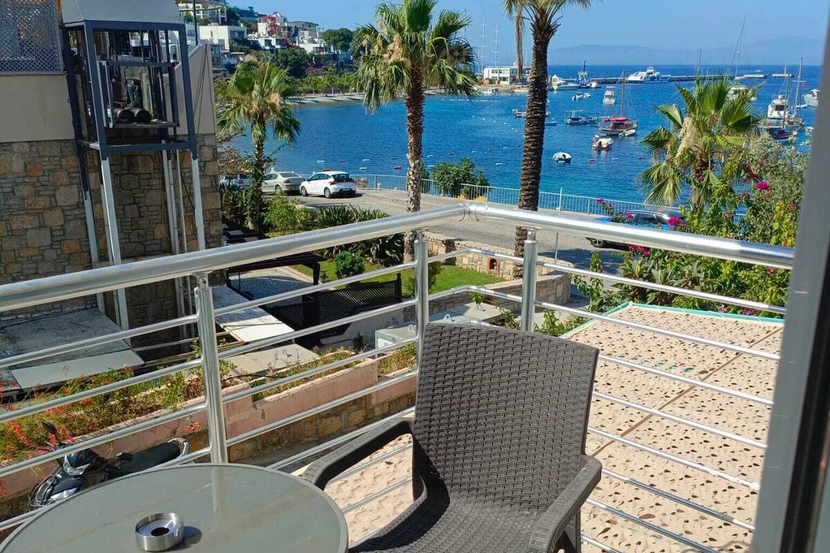 Un balcón con una silla de mimbre y una mesa, con vistas a una marina con barcos y palmeras.
