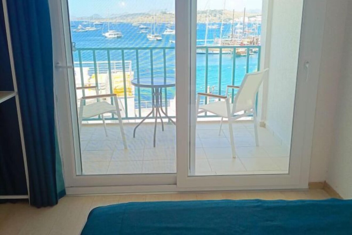 A balcony with a view of the sea and boats, white chairs, and a small table.