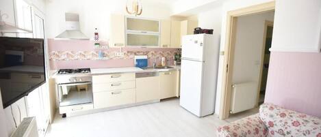 A compact kitchen with white cabinets, a gas stove, a sink, and a refrigerator. There is a floral-patterned sofa in the adjacent living area.