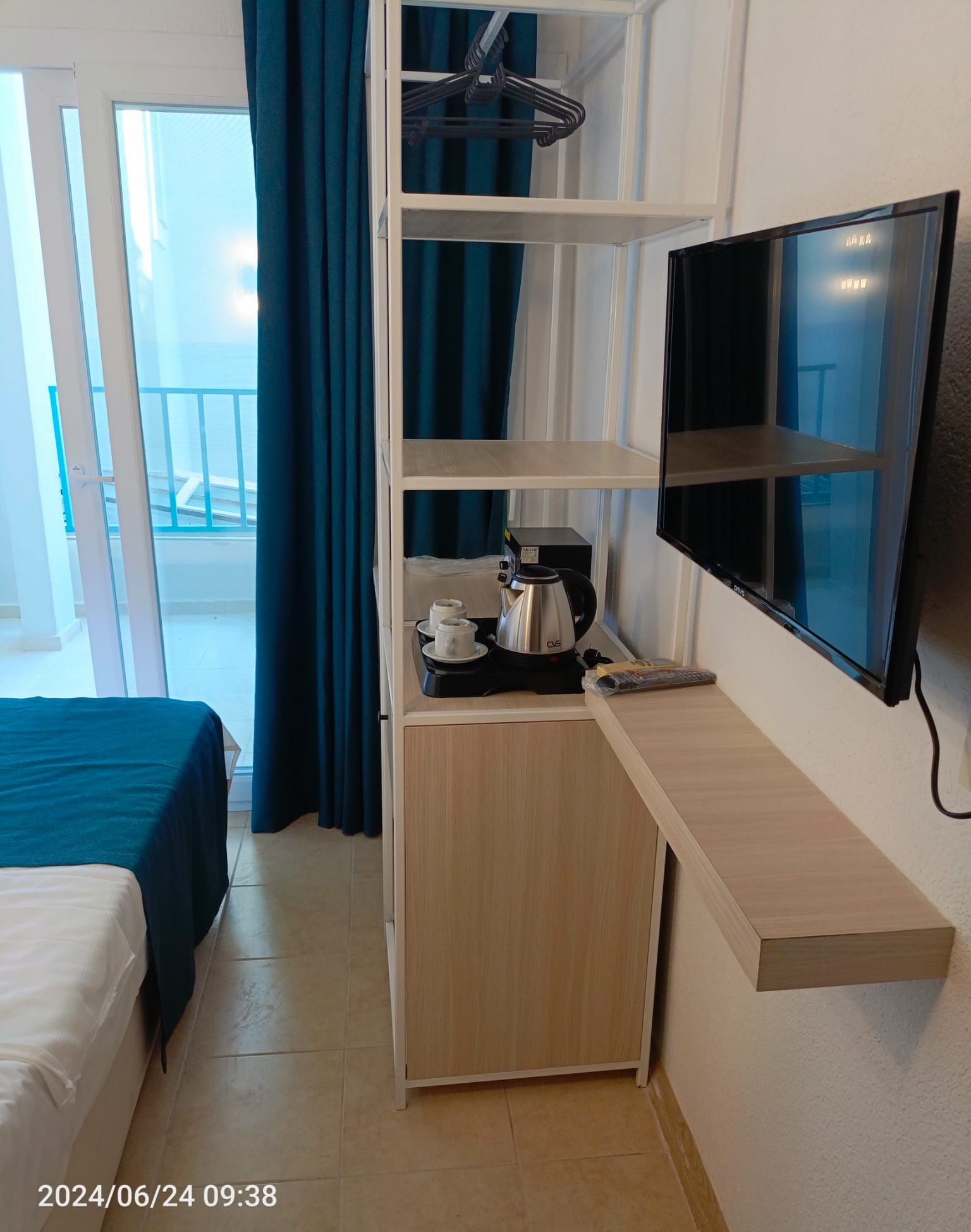 A hotel room with a bed, a desk with a kettle, a TV, and a balcony with blue curtains.