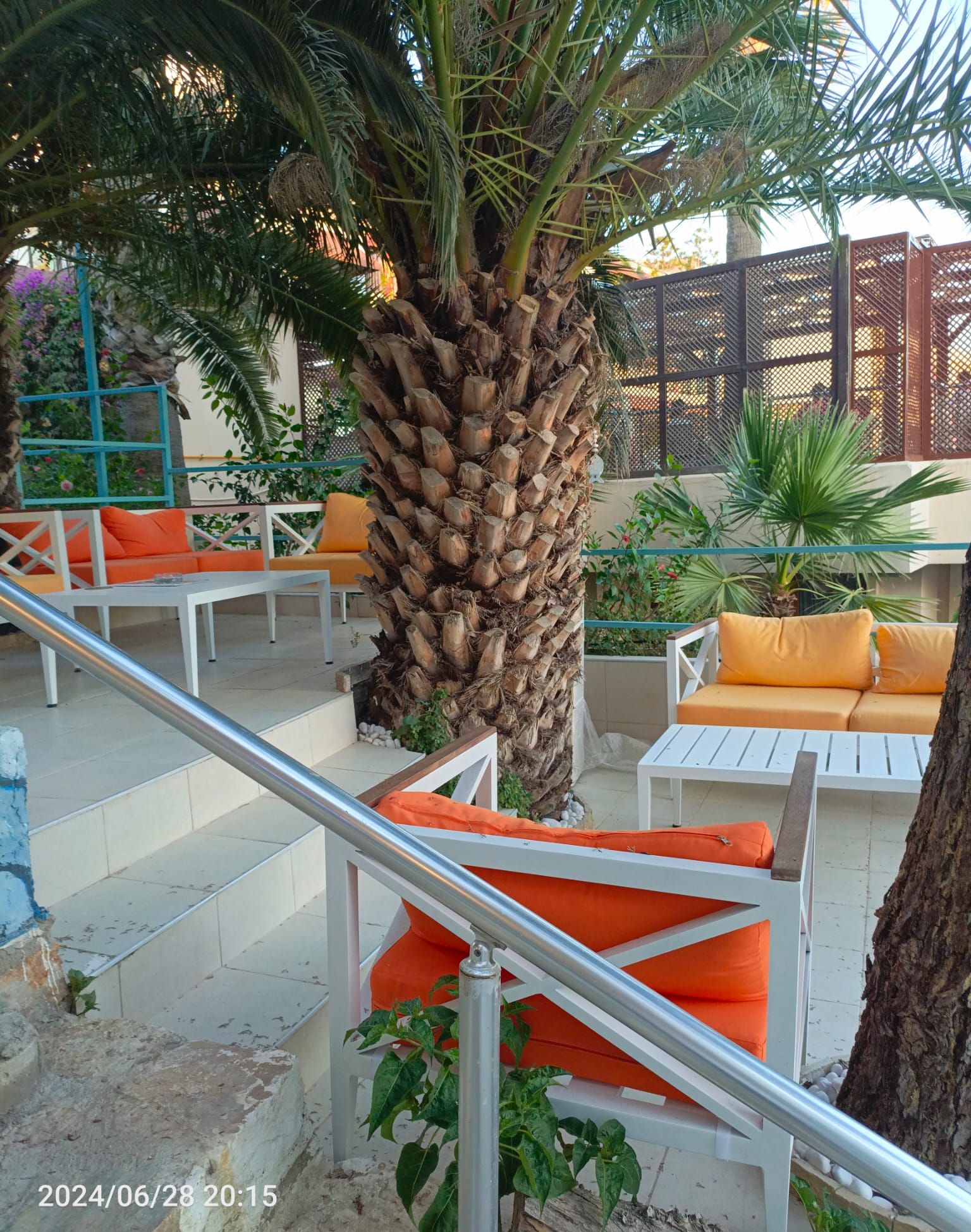 An outdoor seating area with white tables and orange cushions, surrounded by palm trees and a metal railing.