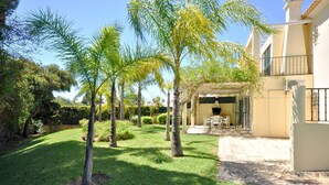 A well-maintained garden with palm trees, a patio area with outdoor furniture, and a building with a beige facade. - Villa in Vilamoura near Falésia Beach (Quarteira)