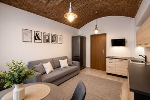 Ein modernes Wohnzimmer mit grauem Sofa, einem kleinen runden Tisch, einem Blumentopf und einer Küchenzeile mit Fernseher und Holzschränken.