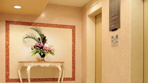 A lobby with a marble floor, a white console table with a floral arrangement, and an elevator with a sign above it. - Hotel Pearl City Morioka (Morioka)