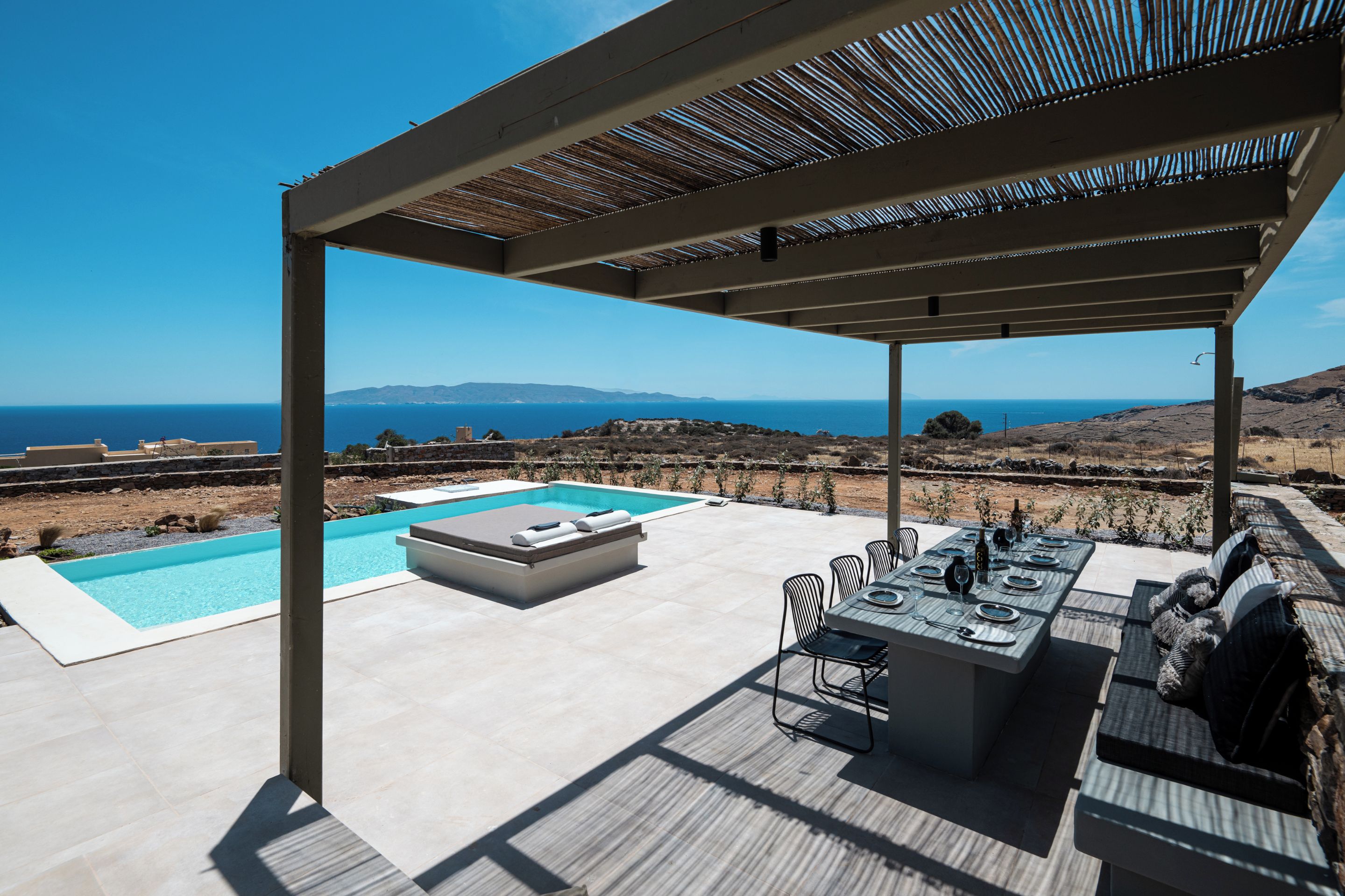 A covered outdoor area with a pool, seating, and a table set for dining, overlooking a scenic landscape.
