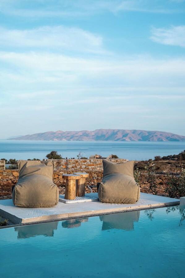 A poolside area with two cushioned chairs and a small wooden table, overlooking a mountainous landscape and the sea. - Onos Luxury Villas Kythnos (Kithnos)