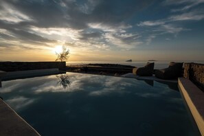 A serene infinity pool with a clear view of the sunset over the ocean, surrounded by stone walls and minimalist seating. - Onos Luxury Villas Kythnos (Kithnos)