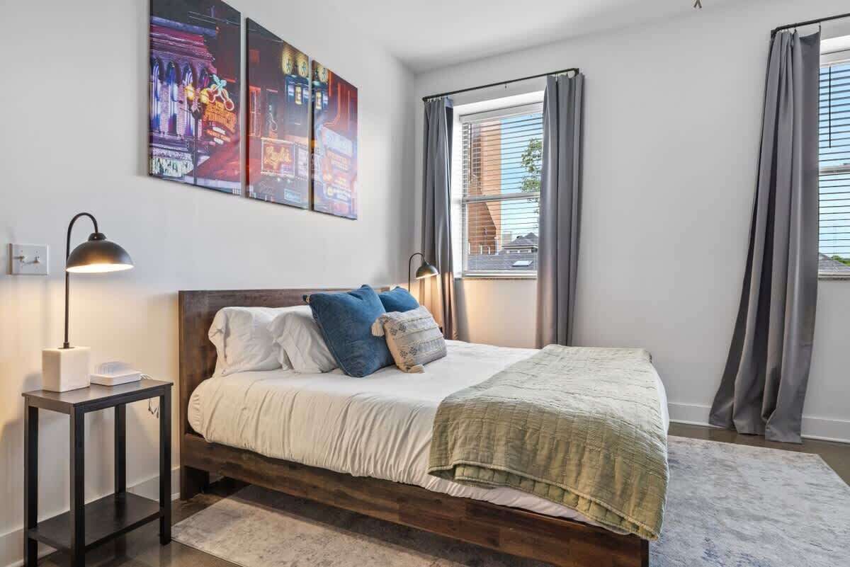 A bedroom with a bed, bedside lamps, a nightstand, a window with curtains, and wall art.