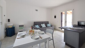 A compact living space with a dining area, a grey sofa with patterned cushions, a small kitchen area, and a television. - Wonderful  villa for 7 guests with WIFI, A/C, TV and patio (Budoni)