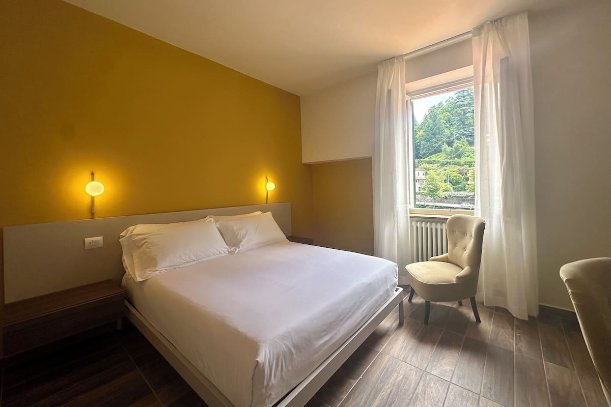 A hotel room with a bed, two wall-mounted lights, a window with curtains, and a chair.
