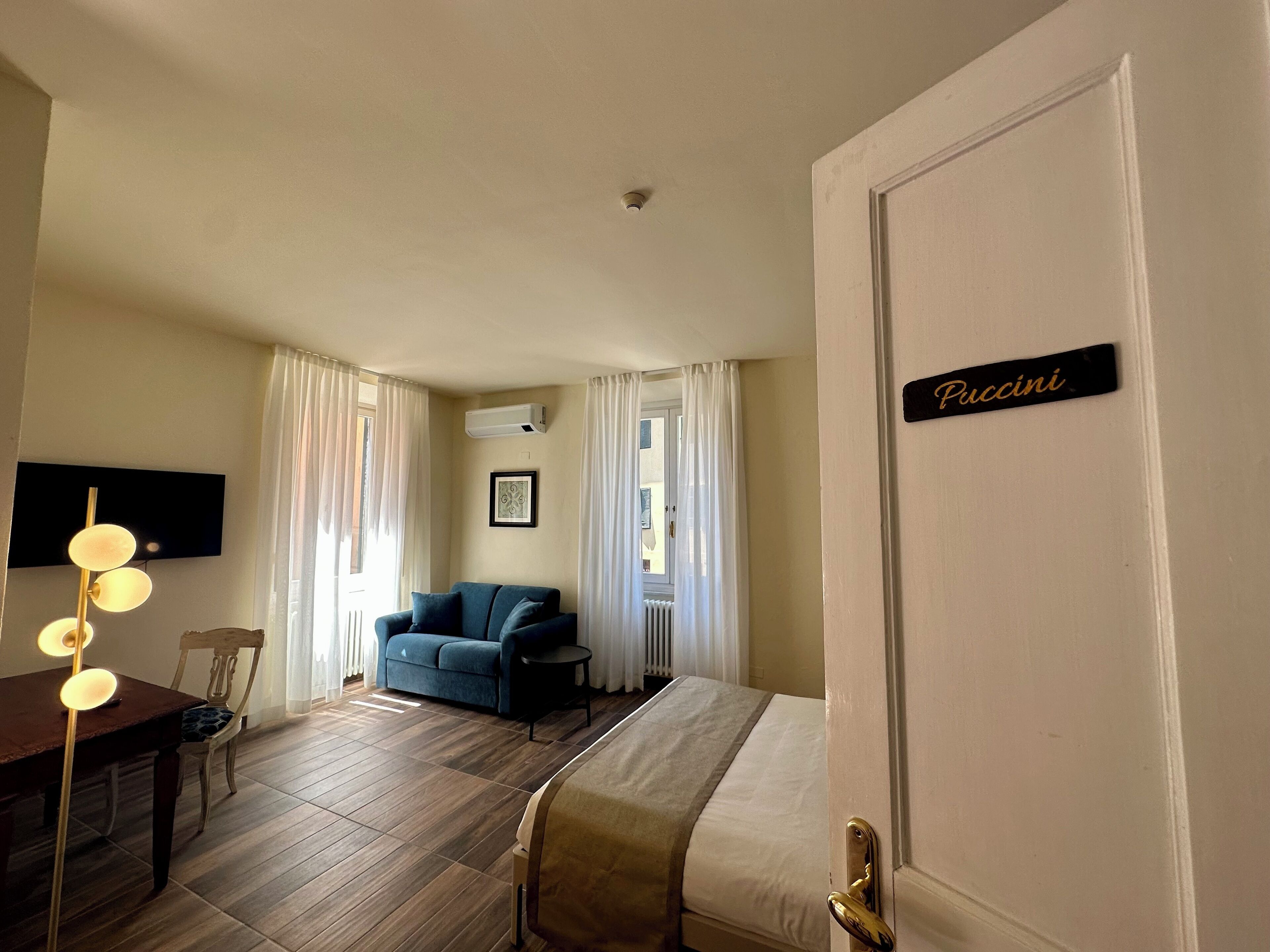A hotel room with a bed, a blue sofa, a wooden dining table with chairs, a TV mounted on the wall, and a sign on the door.