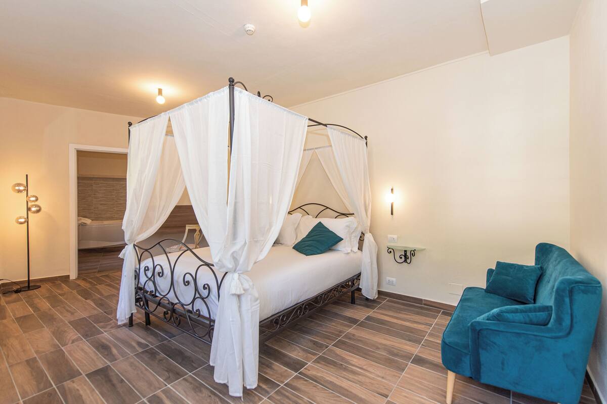 A bedroom with a four-poster bed, a teal armchair, and a nightstand with a lamp.