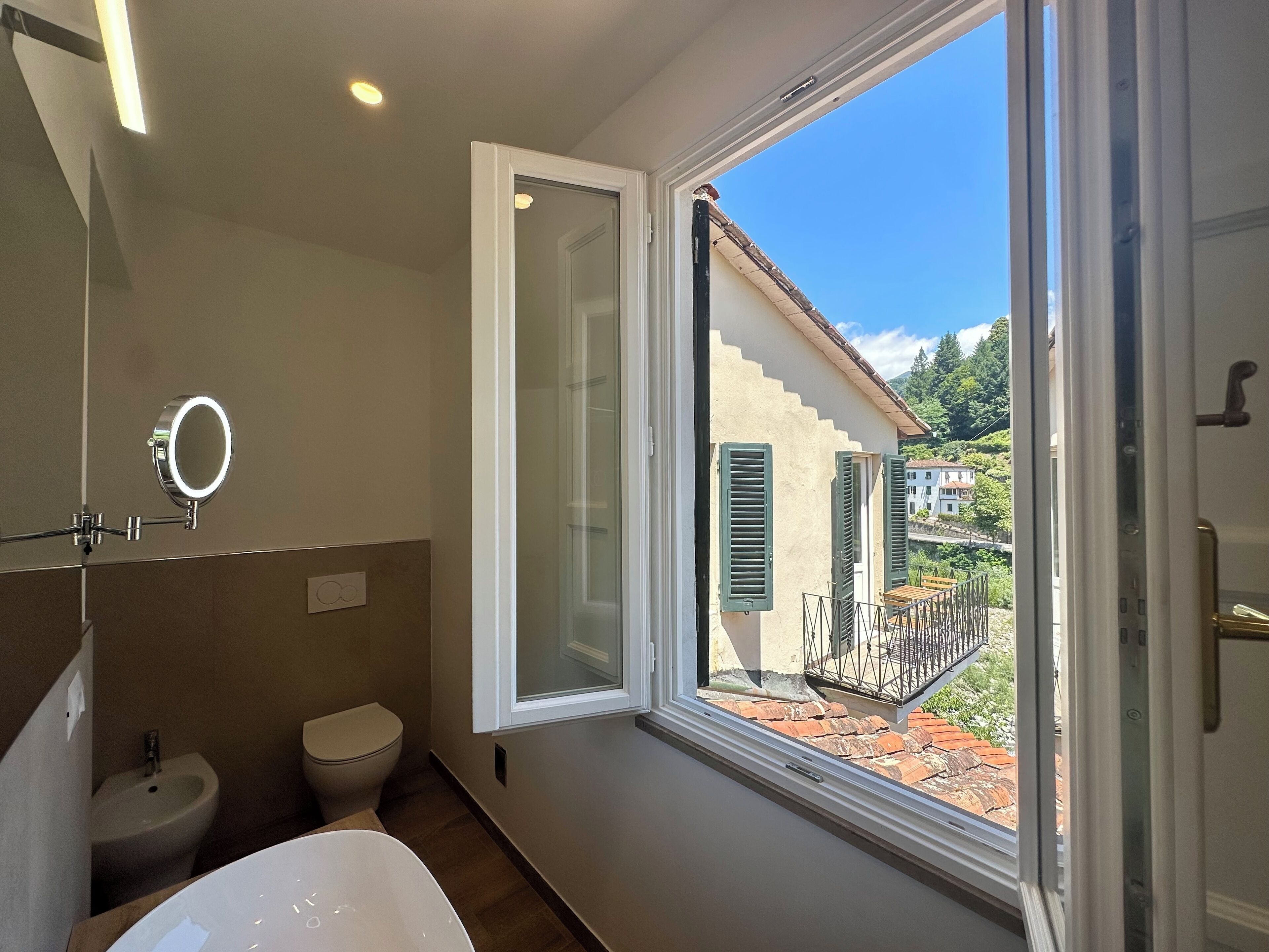 A bathroom with a toilet, bidet, and sink. There is a window with a view of houses and a balcony.