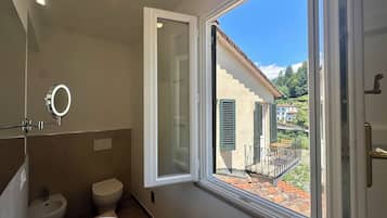 Ein Badezimmer mit Toilette, Bidet und Waschbecken. Ein Fenster bietet Blick auf Häuser und einen Balkon.