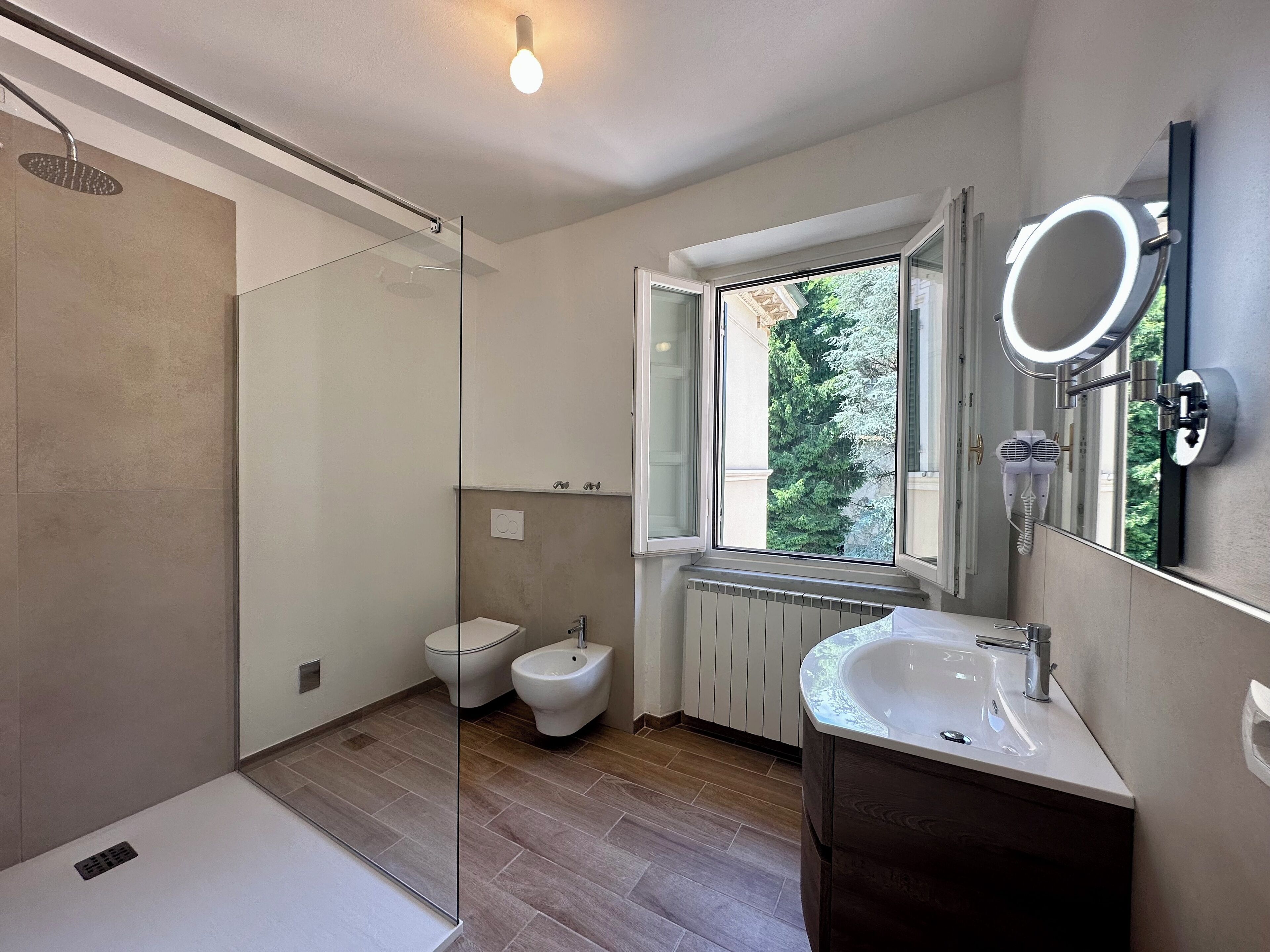A modern bathroom with a glass shower enclosure, a bidet, a sink with a mirror, and a window providing a view of trees.