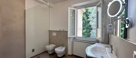 A modern bathroom with a glass shower enclosure, a bidet, a sink with a mirror, and a window providing a view of trees.