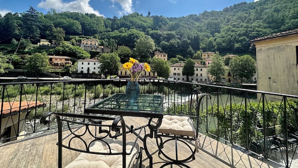 Ein Balkon mit Glastisch und Stühlen, mit Blick auf einen Fluss und ein Dorf mit Gebäuden und Grünflächen.
