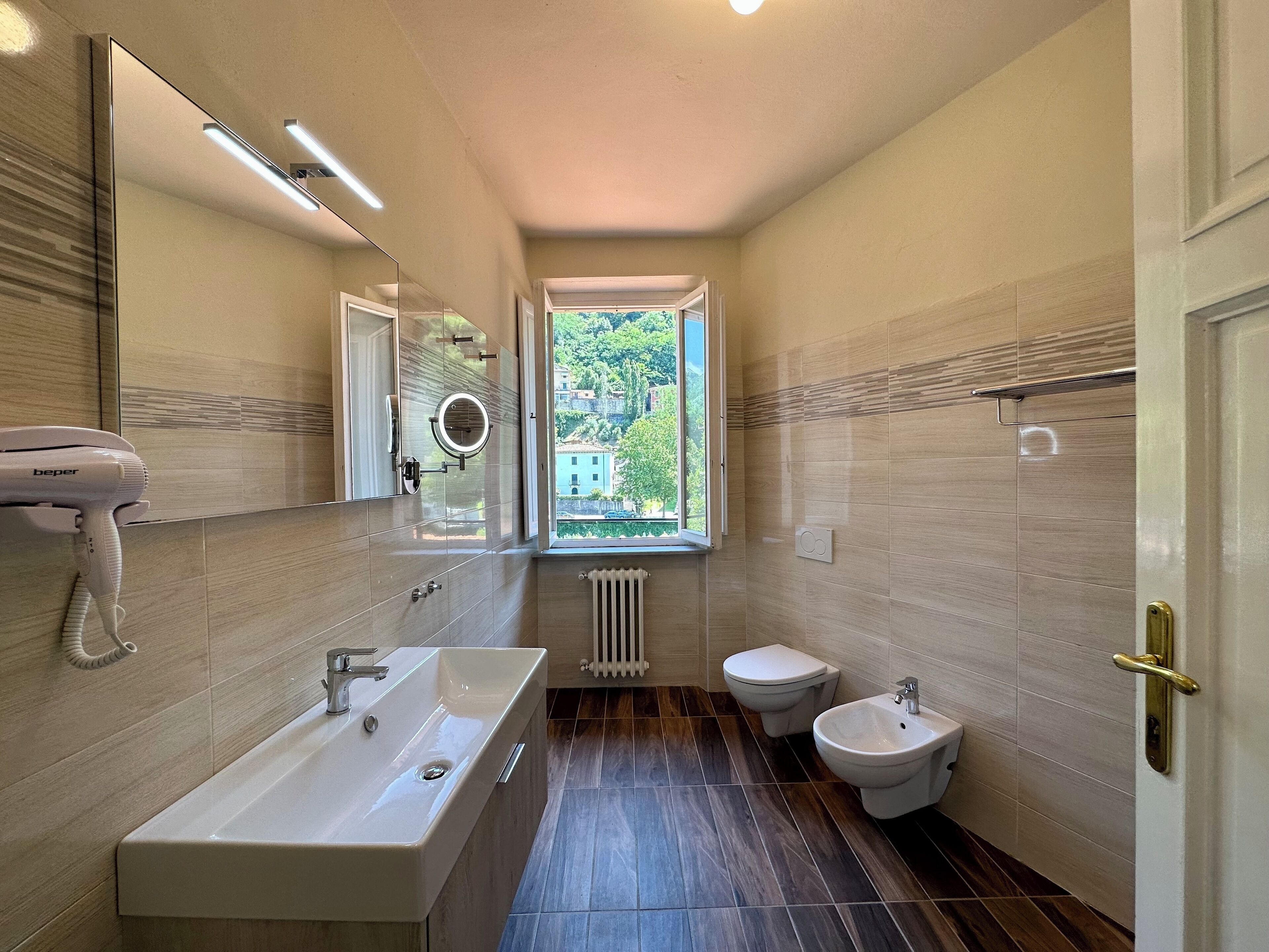A modern bathroom with a large sink, a mirror, a hairdryer, a bidet, and a window providing a view of greenery outside.
