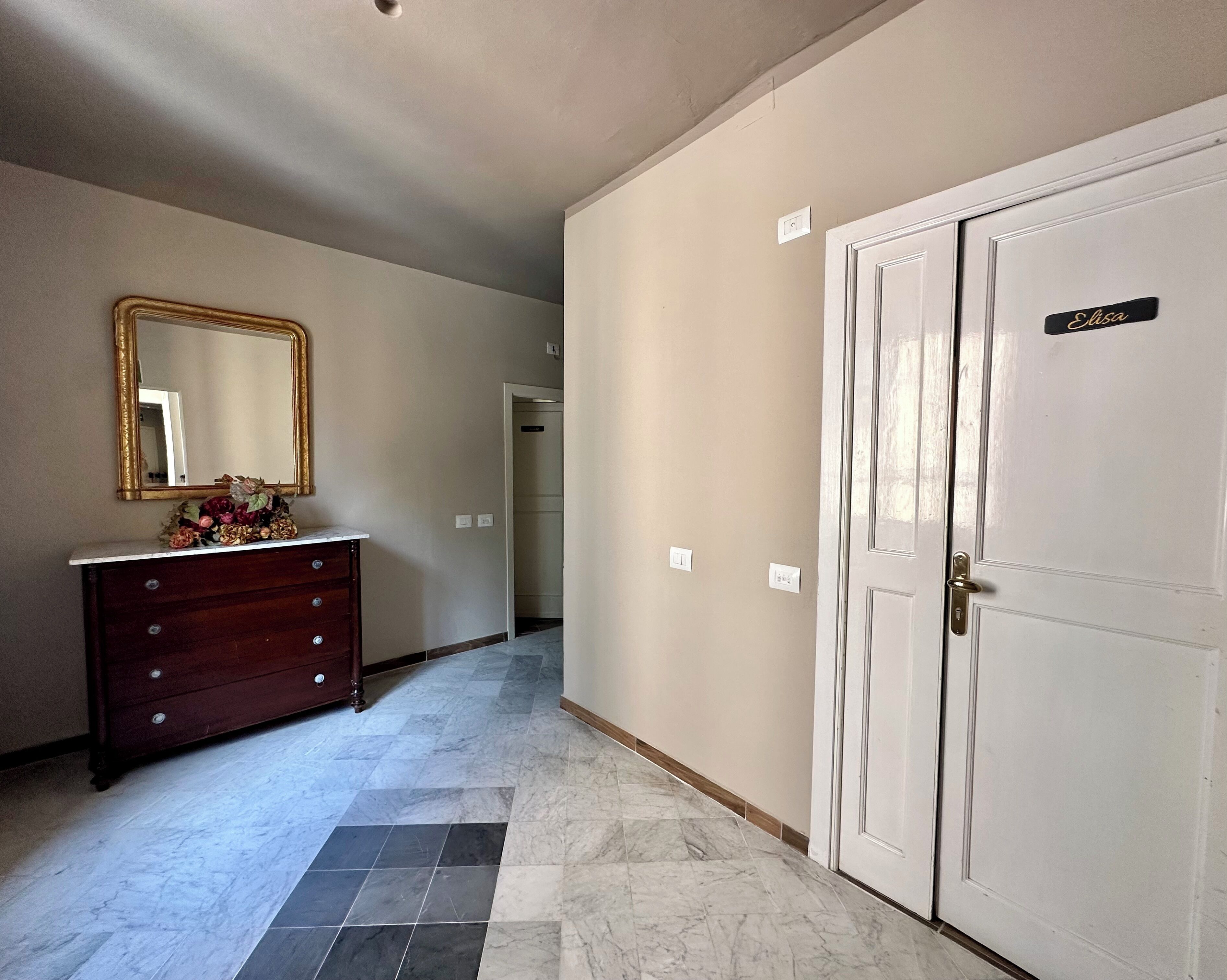 A hallway with a marble floor, a wooden dresser with drawers, a mirror, and a door labeled "Elisa".