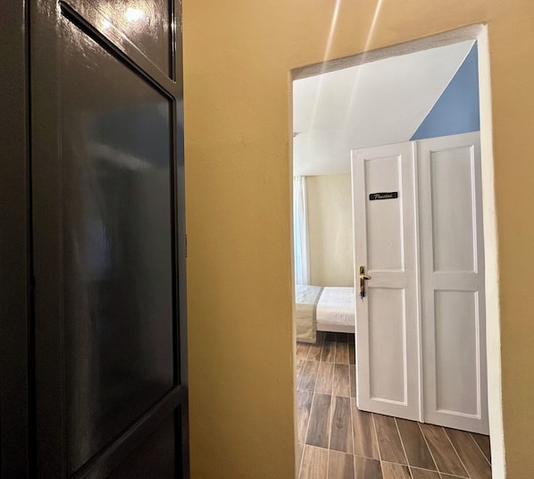 A hallway with a closed dark door on the left and an open white door with a sign reading "Puccini" on the right, leading to a room with a bed and a window. - La Ninfa Hotel (Bagni di Lucca)