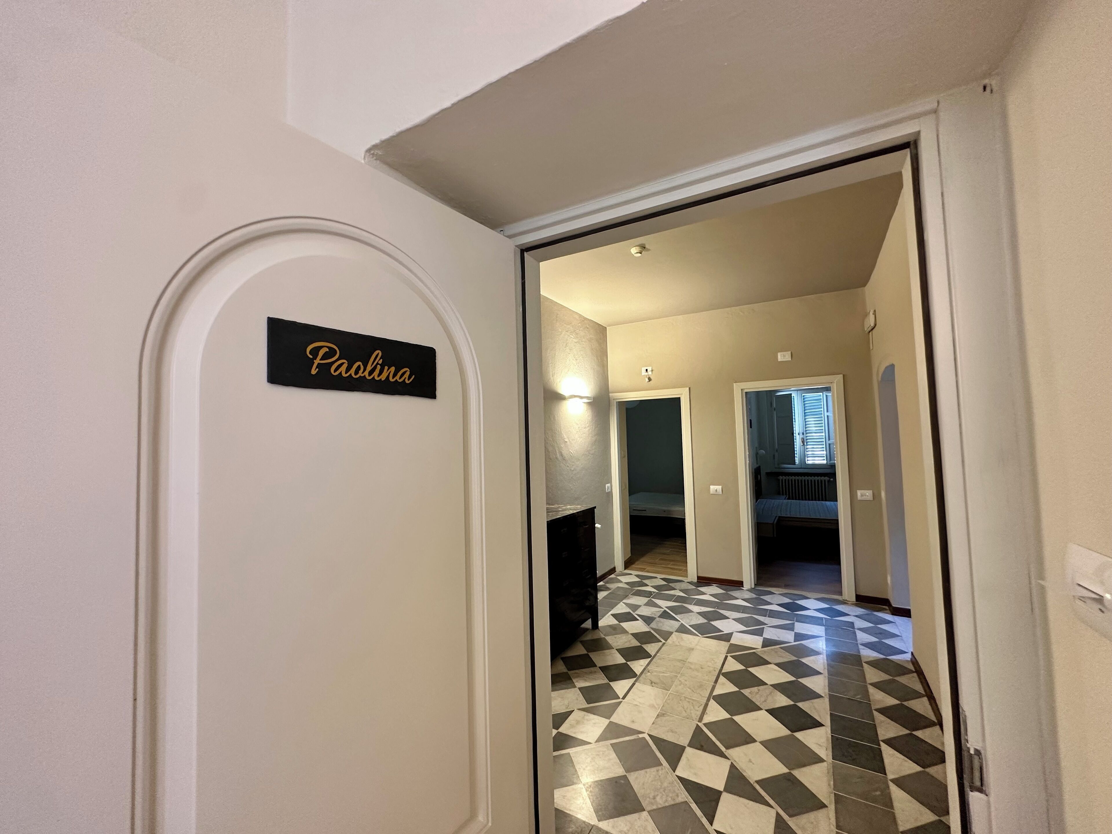 A hallway with a black and white checkered floor, a nameplate reading "Paolina," and a door leading to another room.