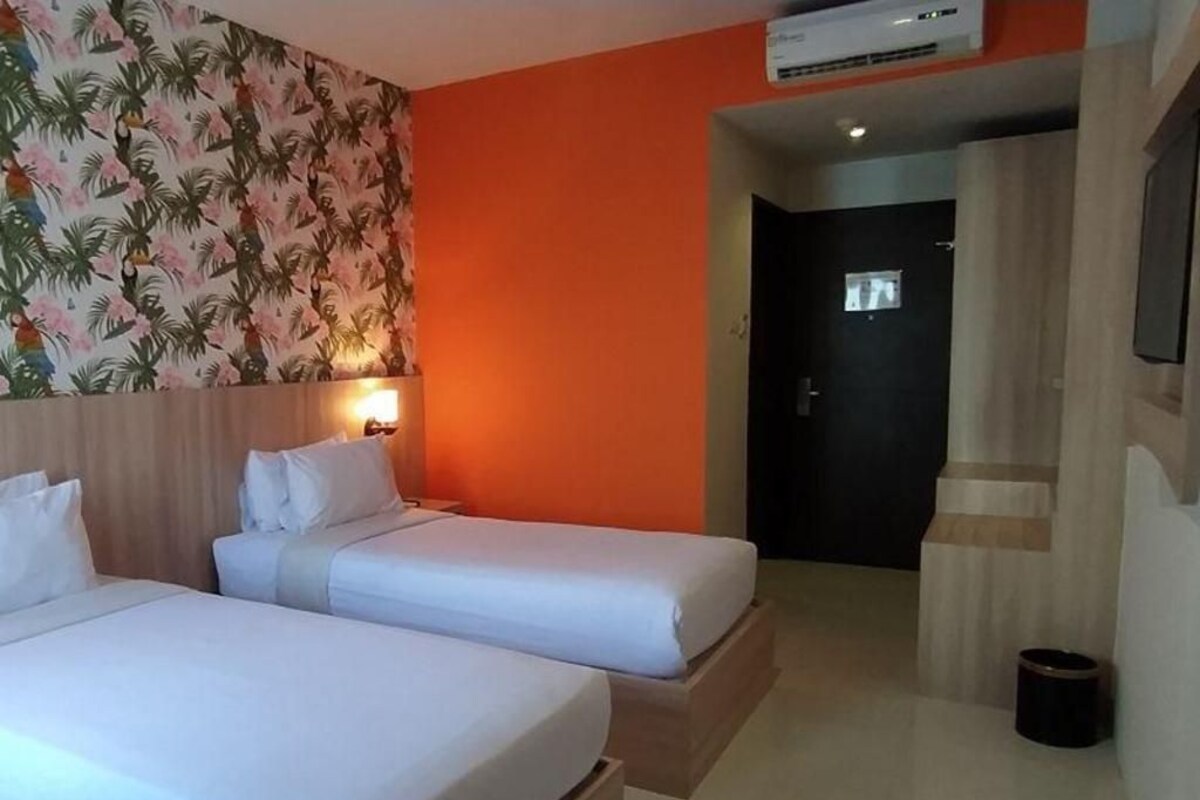 A hotel room with two beds, an orange wall, a tropical wallpaper, and a door leading to another room.