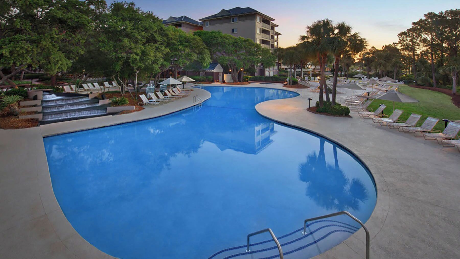A large, curved swimming pool with a clear blue water surface, surrounded by lounge chairs and palm trees.