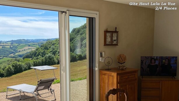 Ein Zimmer mit einer Schiebetür zu einer Terrasse, auf der sich ein Tisch, Stühle und ein kleiner Tisch mit einem dekorativen Gegenstand befinden.