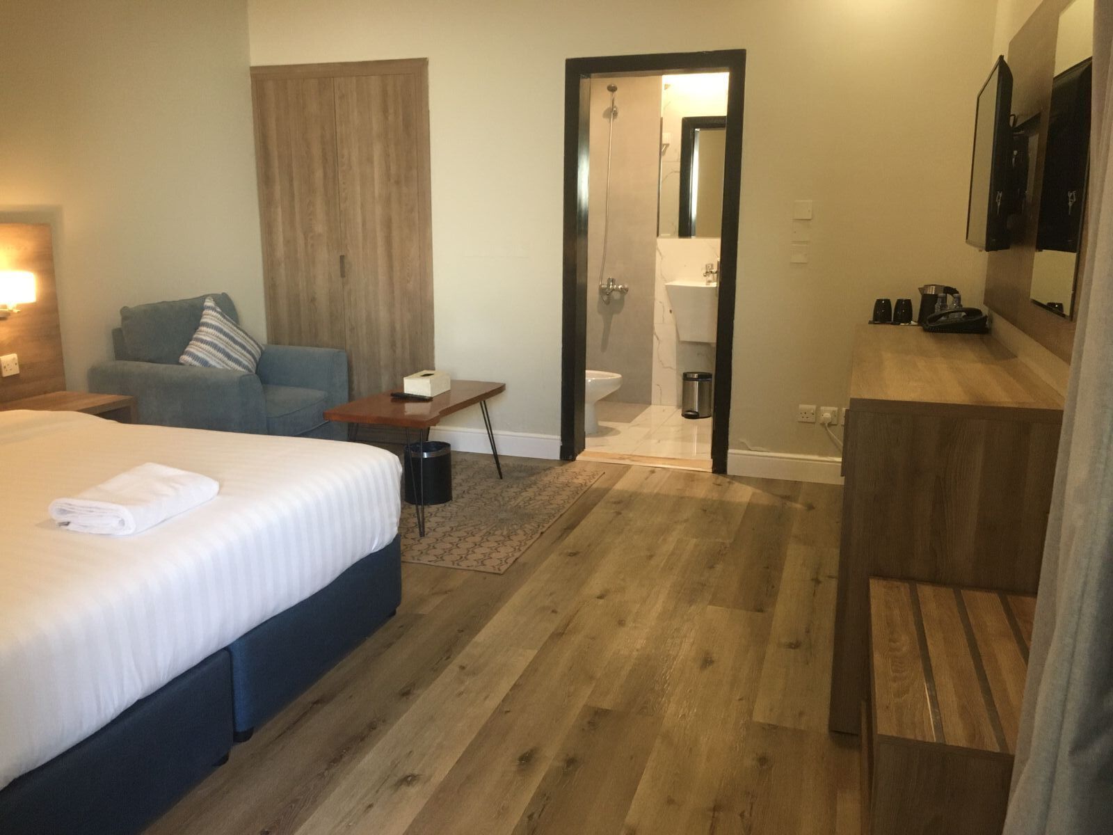 A hotel room with a bed, a sofa, a small table, a desk, a mirror, a shower, and a bathroom.