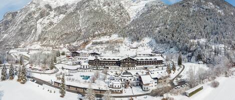 A snow-covered mountain resort with multiple buildings, including a large lodge and guesthouses, surrounded by trees and a clear sky.