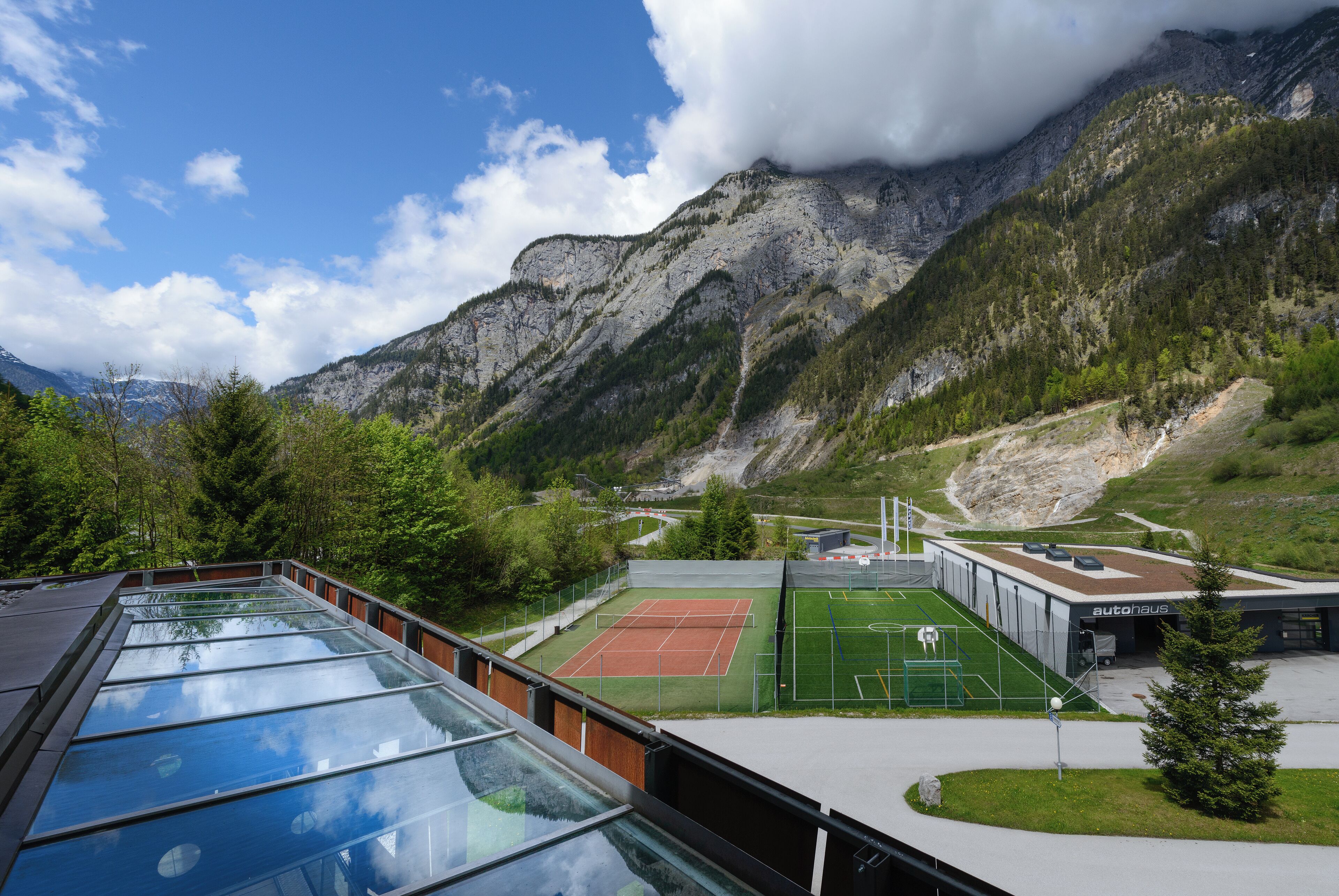 A tennis court with a mountain backdrop, a glass-roofed building, and a parking area.
