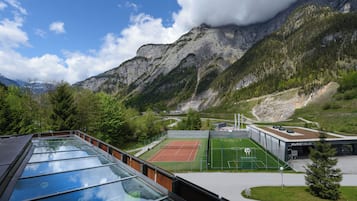 A tennis court with a mountain backdrop, a glass-roofed building, and a parking area.