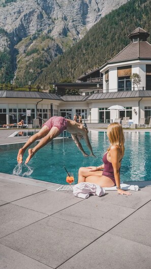 A swimming pool with a person diving into the water, another person sitting by the pool, and a mountain in the background. - Hotel Gut Brandlhof (Saalfelden am Steinernen Meer)