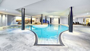 Un'ampia piscina interna con pavimento piastrellato, circondata da lettini e una zona relax con vista su un paesaggio montuoso.