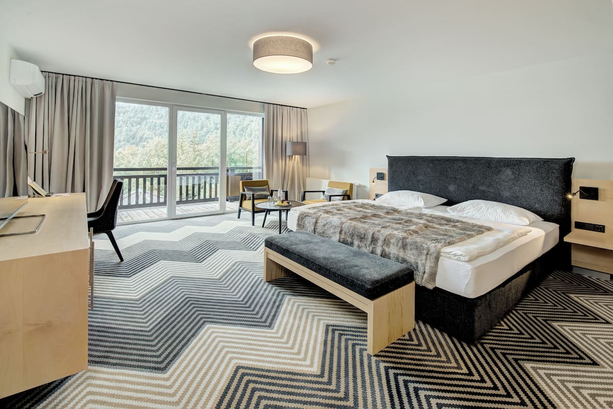 A modern hotel room with a large bed, a desk, a chair, and a balcony with a view of the forest.