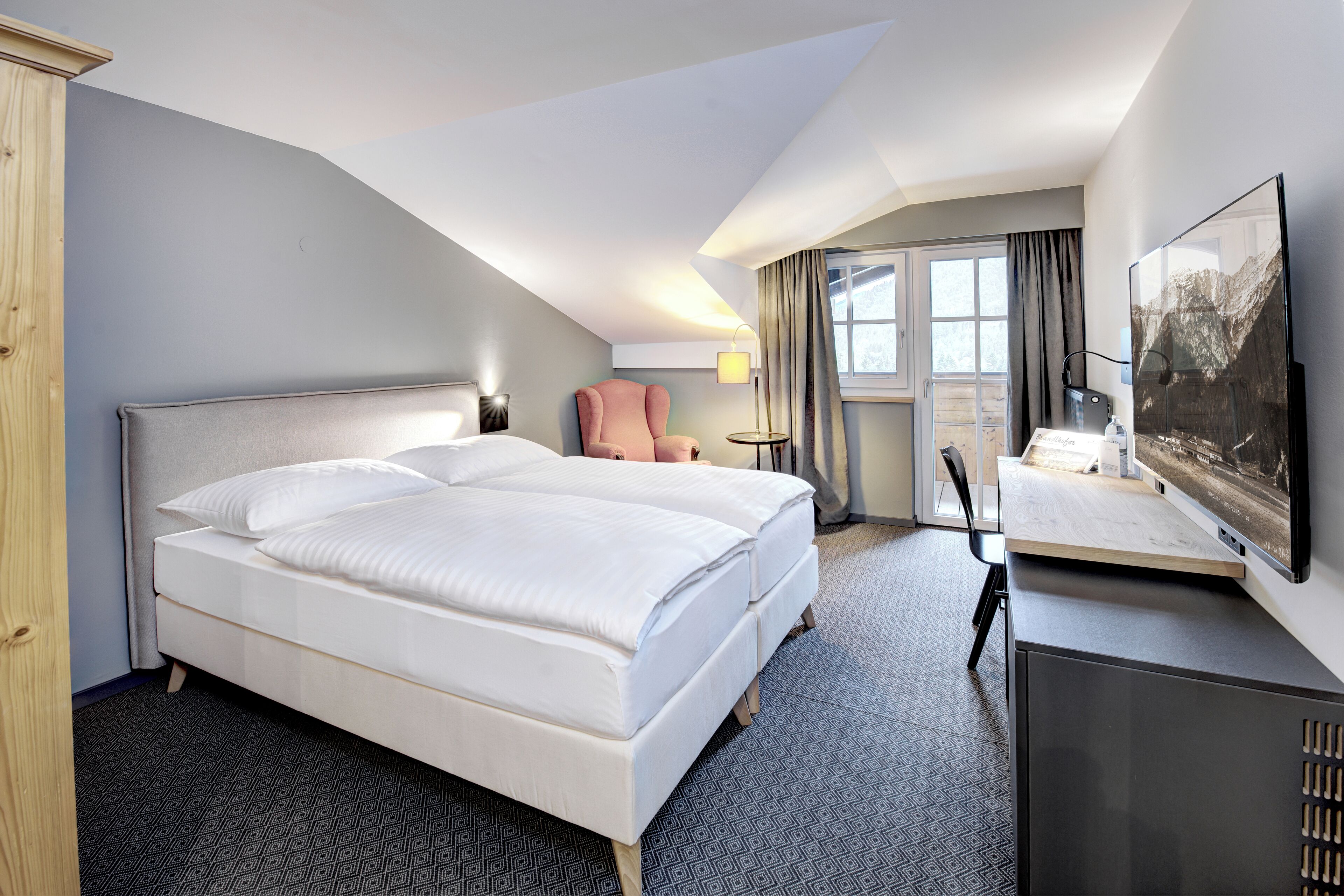Ein Hotelzimmer mit einem großen Bett, einem Schreibtisch, einem Sessel, einem Fernseher und einem Fenster mit Vorhängen.
