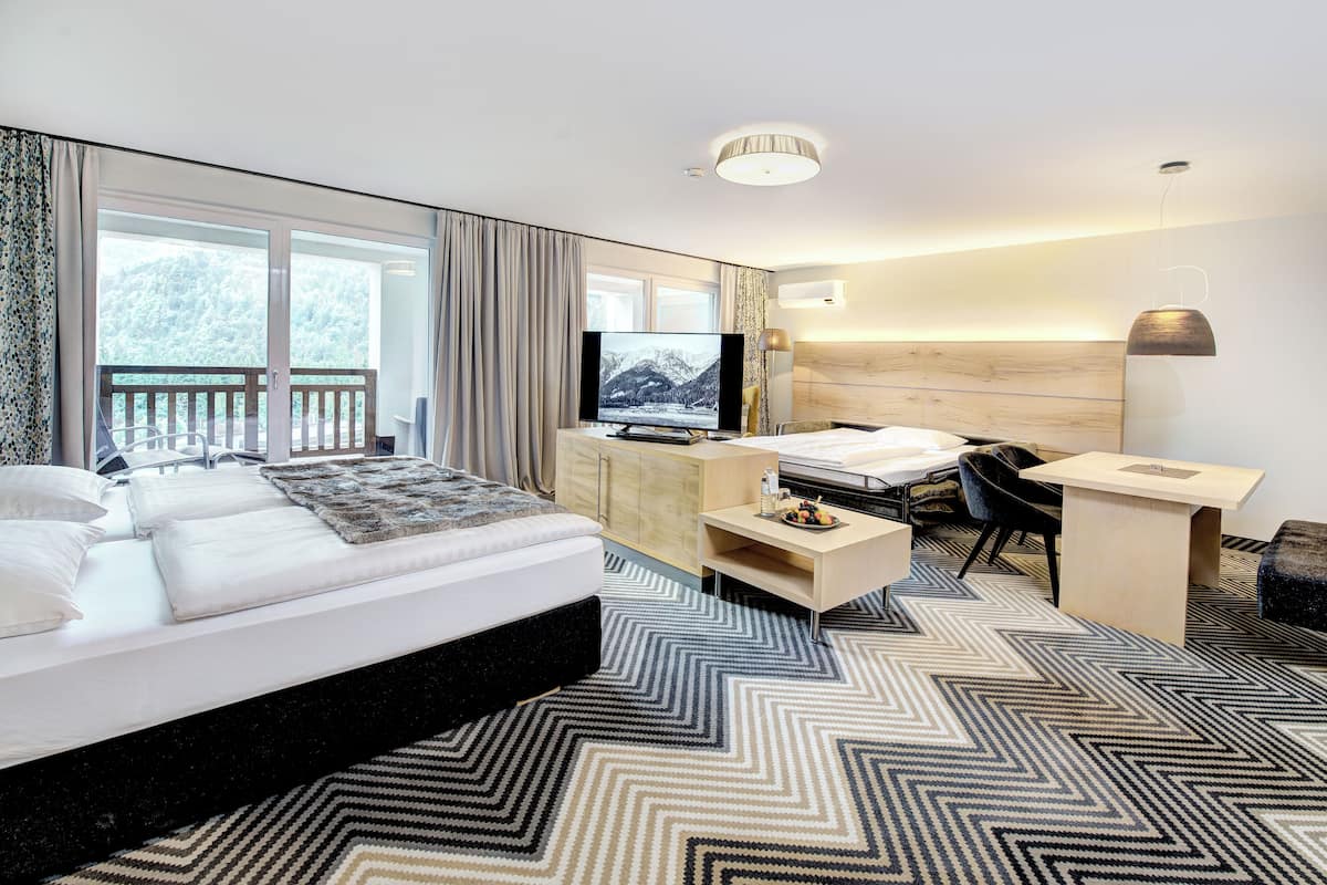 A modern hotel room with a large bed, a desk, a chair, a television, and a view of a balcony with a wooden railing.