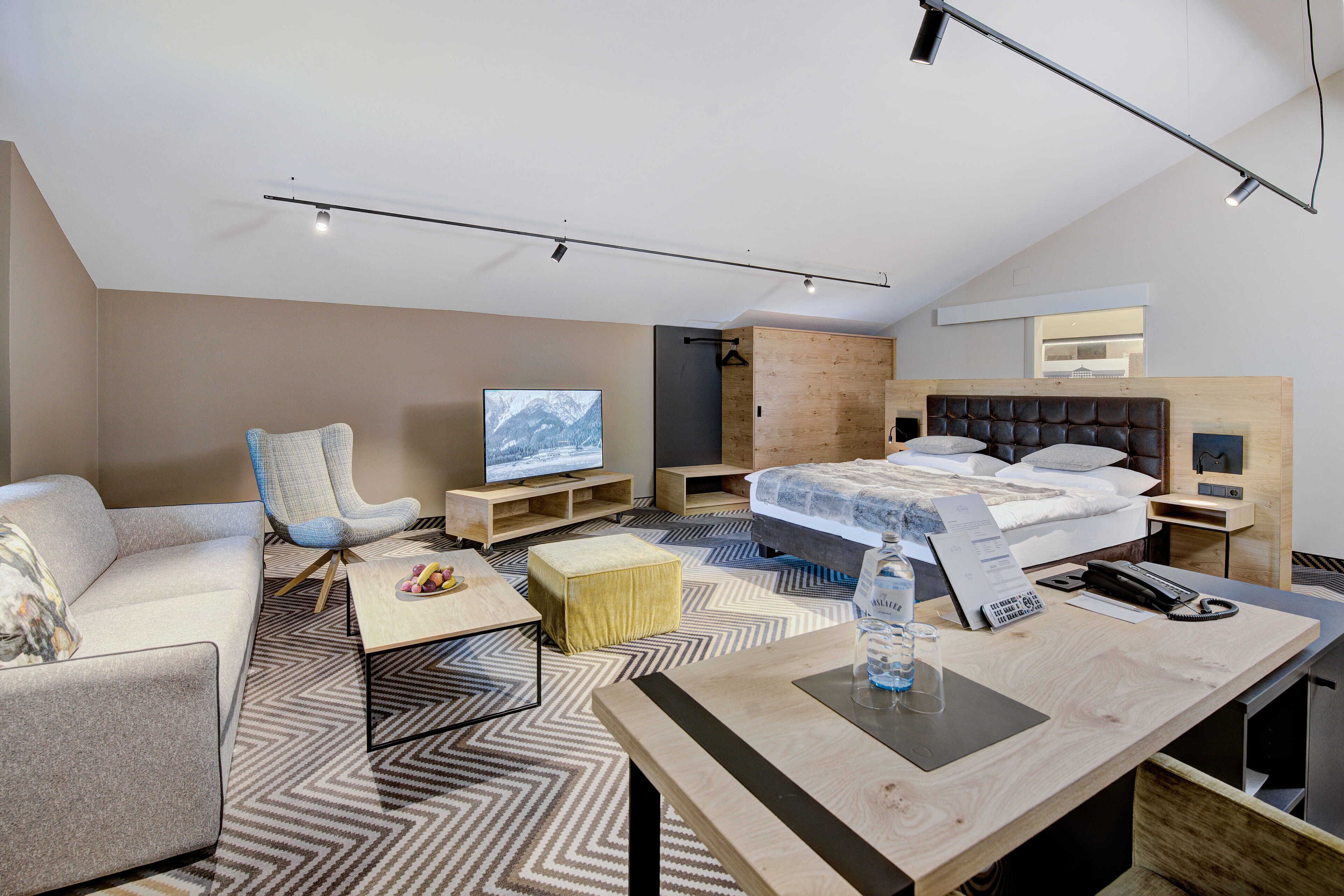 A modern hotel room with a sofa, armchair, wooden bed, and a TV.