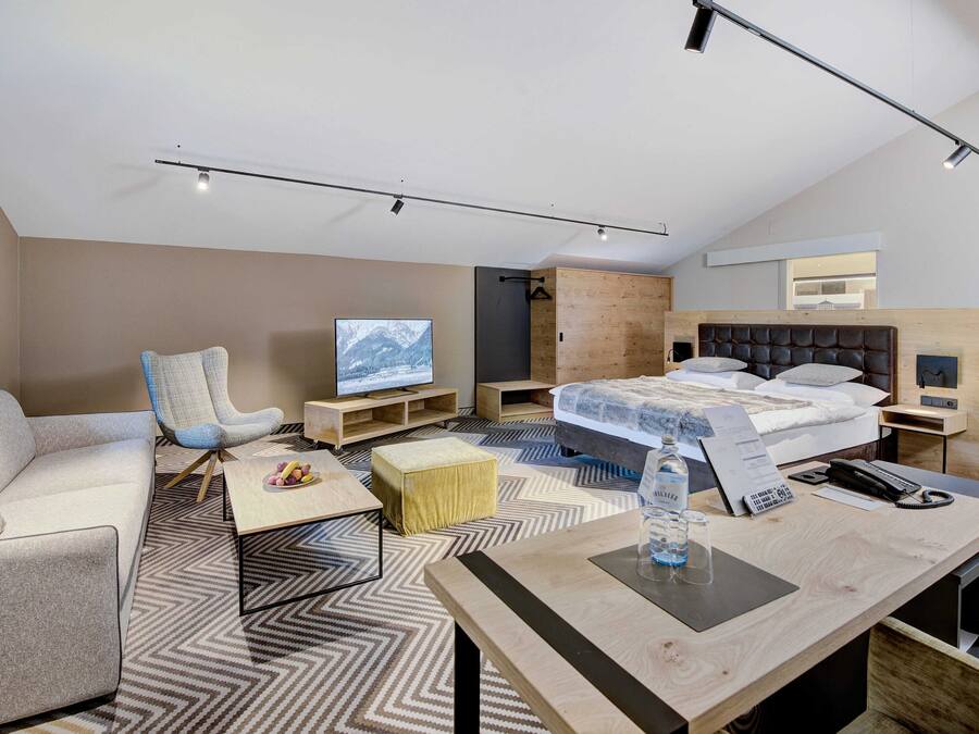 A modern hotel room with a sofa, armchair, wooden bed, and a TV.