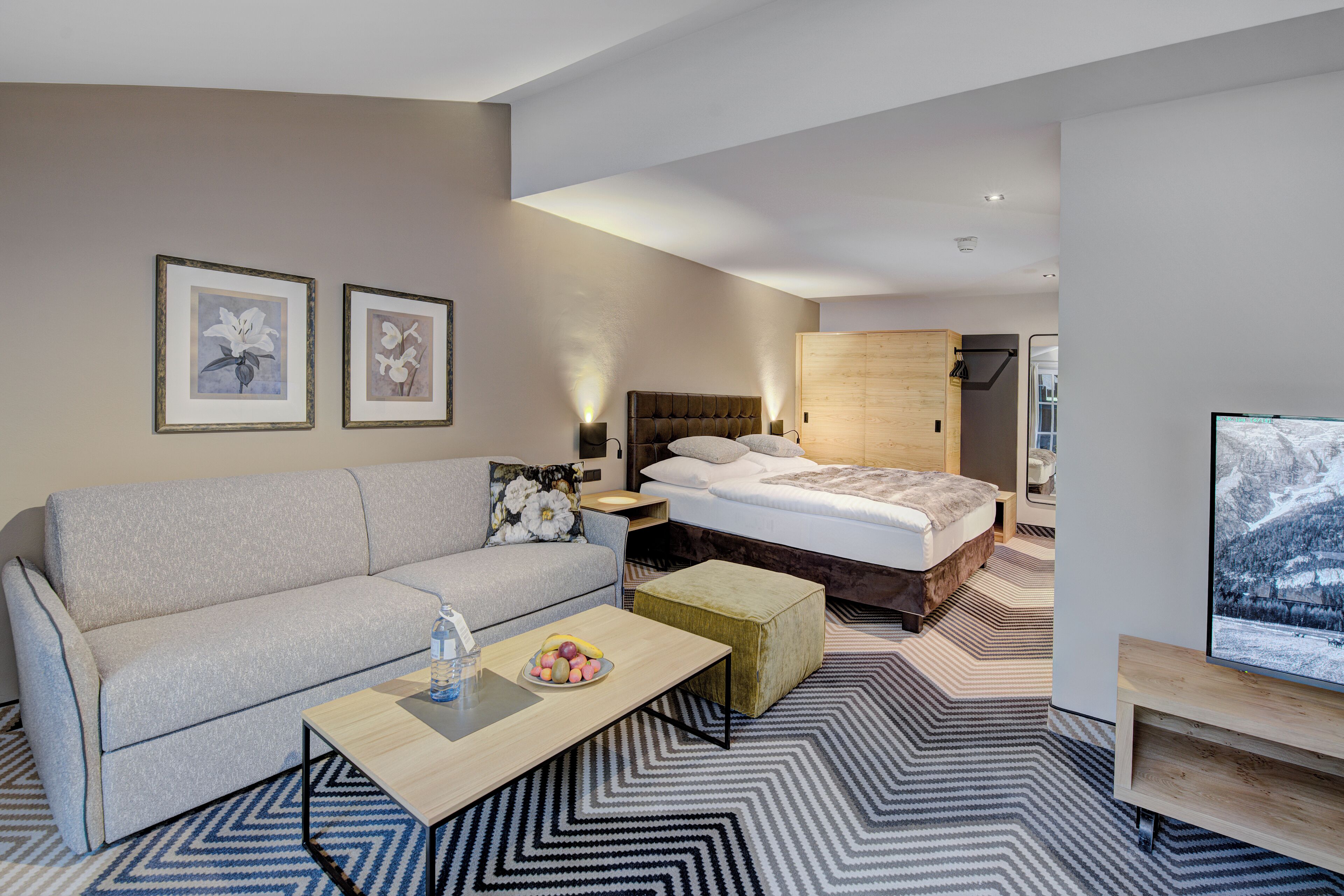 A modern hotel room with a bed, a sofa, a coffee table, and a TV.