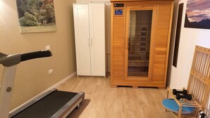 Une pièce équipée d’un tapis de course, d’un sauna en bois et d’un petit espace d’entraînement avec des haltères.