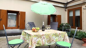 Une terrasse extérieure avec une table recouverte d’une nappe à motifs floraux, deux chaises vertes, une bouteille de vin et un saladier de fruits.