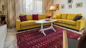 Een woonkamer met een gele bank, een glazen salontafel en een rood tapijt met patroon.