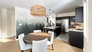 Een moderne keuken met een eetgedeelte, voorzien van een groot schiereiland, hanglampen en uitzicht op een bos.
